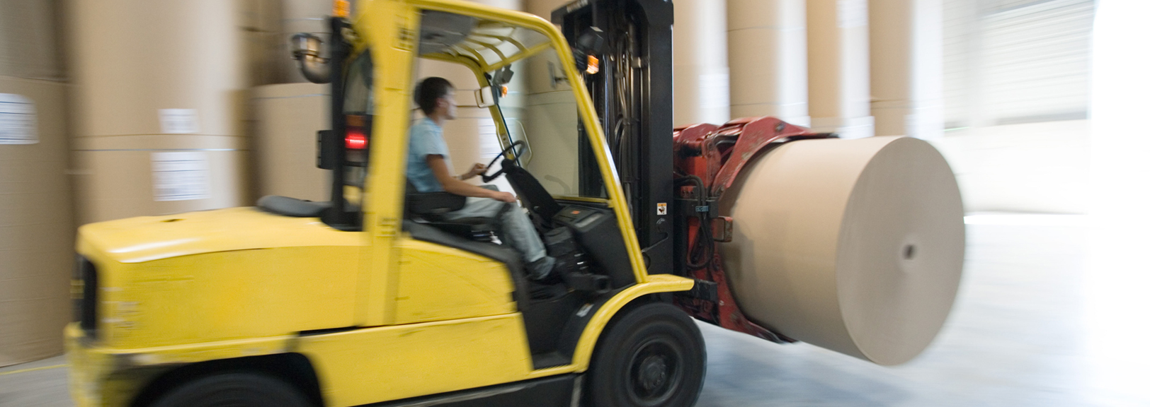 Yellow forklift moving a large paper roll at speed in a warehouse